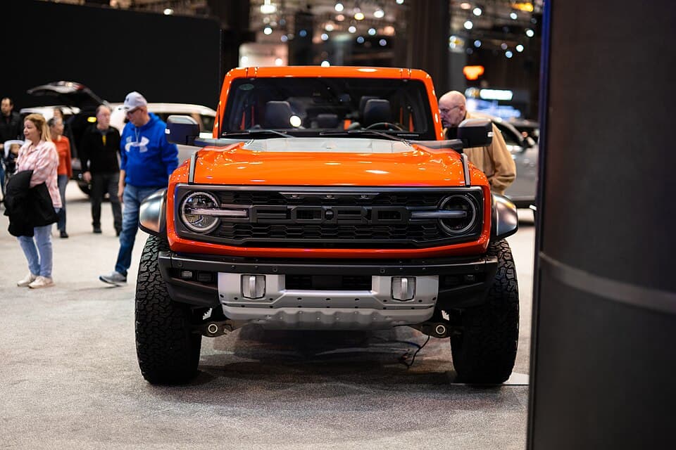 Code Orange Bronco Raptor with the roof panels off at the 2023 Cleveland Auto Show, shot head-on — the widened track and 37-inch tyres are obvious from this angle.