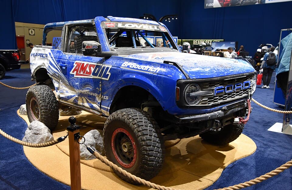 Race-prepped Bronco Raptor in AMSOIL/BFGoodrich livery, built for the King of the Hammers 4600 class and shown on a display stand — caged, stripped and on beadlocks. Not a stock truck.