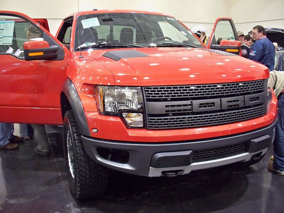 2010 F-150 SVT Raptor on an indoor auto-show floor with the doors open, shot head-on so you can see the full-width FORD block grille and the extra track width over a standard F-150.