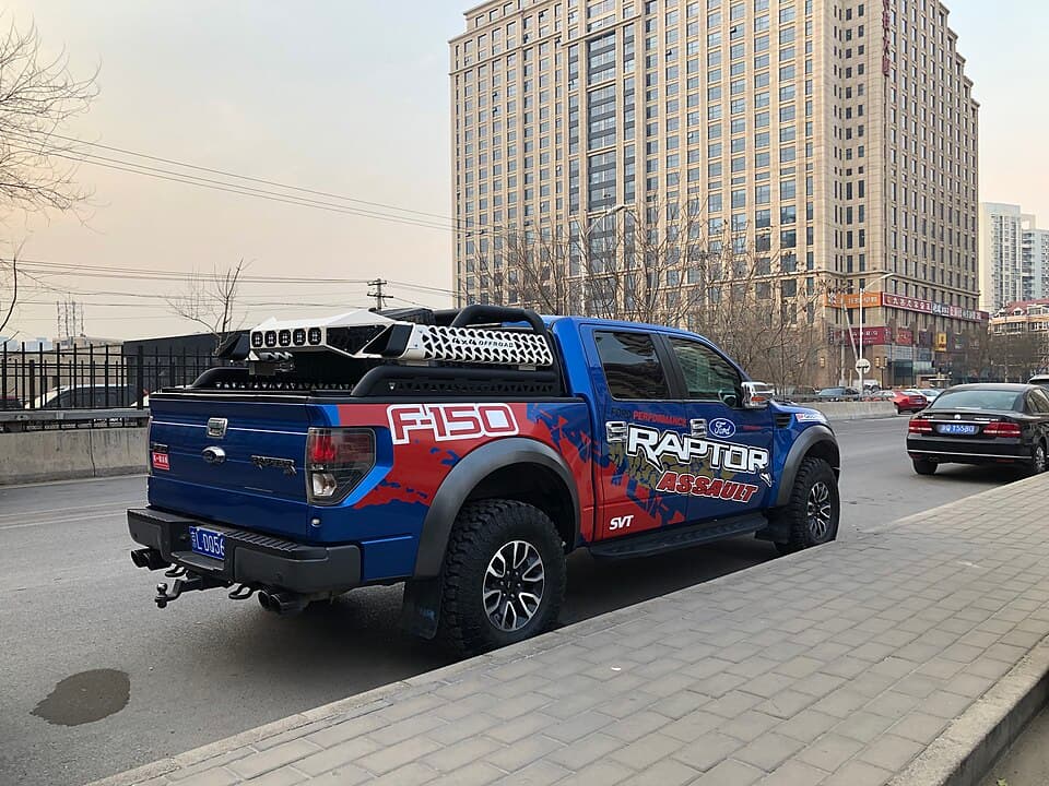 Blue first-gen SVT Raptor with F150 RAPTOR bedside graphics and a bed rack, rear three-quarter on a Beijing street.
