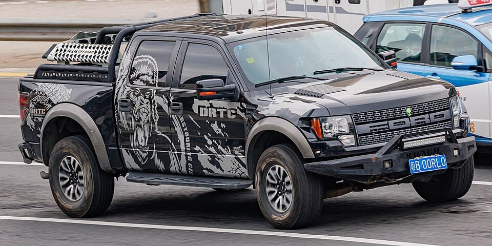 Black first-gen SVT Raptor in DRTC off-road-club livery with a light bar and bed rack, side profile on a street in China.