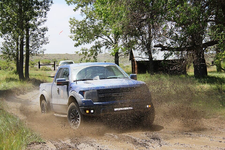 Blue first-gen SVT Raptor SuperCab throwing a rooster tail of mud down a two-track outside Fernley, Nevada — the internal-bypass Fox shocks doing exactly what they were built for.