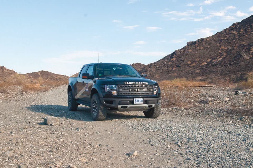 Black first-gen Raptor in RANGE WARDEN livery on a gravel wash in the Barry M. Goldwater Range, Arizona — a US military patrol truck, front three-quarter.