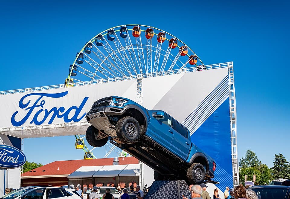 Blue second-gen Raptor tipped up on a display ramp at the Minnesota State Fair with the Ferris wheel behind it — a clean look at the ground clearance and approach angle.