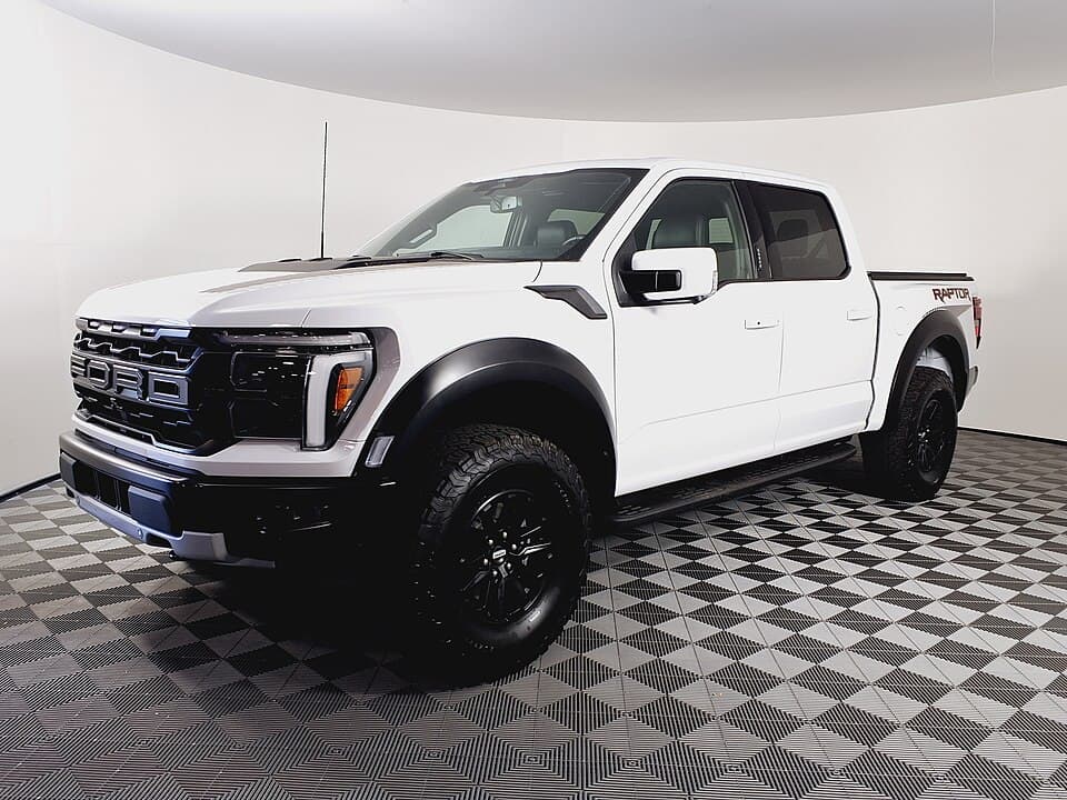 White 2024 Raptor in a dealer showroom, front three-quarter — the refreshed front end with the reprofiled grille and headlights.