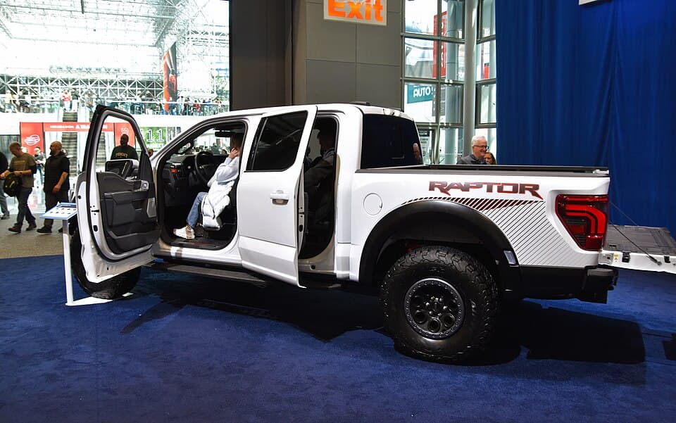 White third-gen Raptor on the stand at the 2026 New York International Auto Show with the doors open, side-on, red RAPTOR bedside graphic visible.
