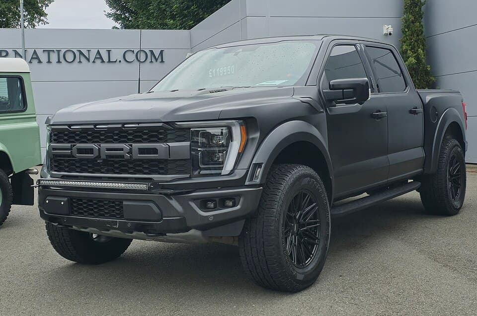 Satin-black third-gen Raptor on a UK dealer forecourt, front three-quarter with aftermarket wheels and a light bar in the lower bumper.